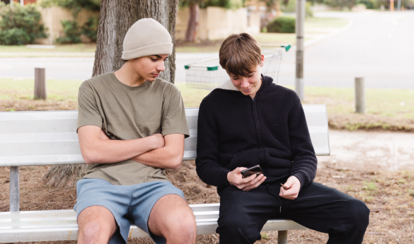 A teenage boy showing another teenage boy his phone on a bench