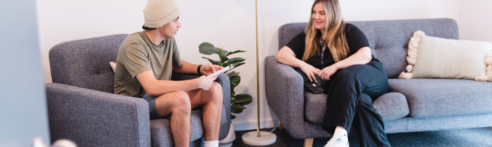A young person and a Youth Futures Djinda staff member sit and chat in a comfortable lounge setting, with the young person holding a folder.