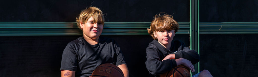Two young students sit together outdoors holding basketballs, lit by warm afternoon sun against a dark background.